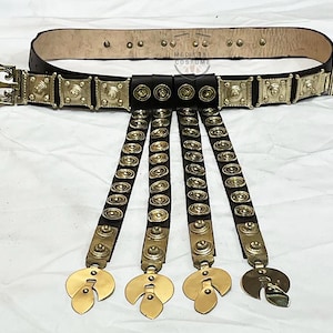 May include: A brown leather belt with gold metal accents and five hanging straps with gold metal discs. The belt has a buckle with a decorative design.