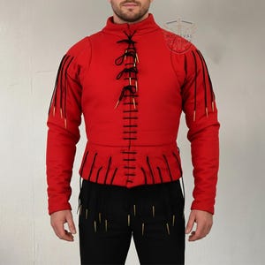 Könnte beinhalten: Ein rotes mittelalterliches Wams mit schwarzer Schnürung und goldfarbenen Akzenten. Die Jacke mit hohem Kragen hat dekorative schwarze Fransen und goldfarbene Verzierungen. Das Kleidungsstück wird mit einer schwarzen Hose kombiniert, die ebenfalls mit goldenen Akzenten verziert ist.