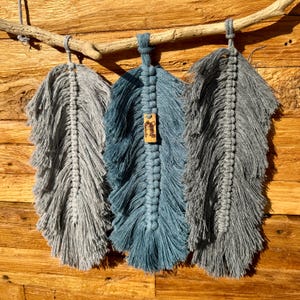 May include: Three macrame feather wall hangings in shades of grey and blue, hanging from a driftwood branch. The centre feather has a small wooden tag with the word "Love" written on it.