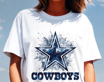 Op de afbeelding: Wit T-shirt met een blauw en wit sterontwerp, met een silhouet van een stadsgezicht. Het woord "COWBOYS" is in blauw onder de ster gedrukt. Het ontwerp heeft een aquareleffect.