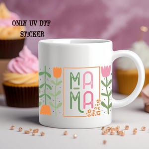 A white mug with a colorful design featuring the word "MAMA" and floral elements, placed on a table with cupcakes in the background.