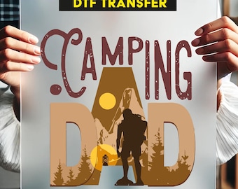 May include: Ready to press DTF transfer featuring the text "Camping Dad" in red and brown. The design depicts a silhouette of a person hiking in a mountain landscape with trees and a sunset.