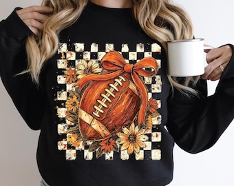 May include: Black sweatshirt featuring a football design with a large orange bow and floral accents. The football is brown with white stitching. The background is a distressed black and white checkered pattern. The design is centered on the front of the shirt.
