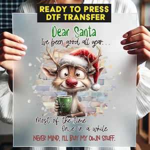 May include: A clear sheet with a cartoon reindeer wearing a Santa hat and holding a green mug. Text reads "Dear Santa, I've been good all year... Most of the time... Once in a while... NEVER MIND, I'LL BUY MY OWN STUFF." The top text says "READY TO PRESS DTF TRANSFER."