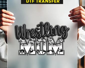 以下が含まれることがあります： 「Wrestling Mom」というテキストが、黒と白の迷彩柄で書かれた、プレス可能なDTF転写シート。テキストはレスラーのシルエットで囲まれています。
