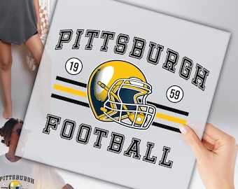 Peut inclure: Graphique blanc avec les mots "PITTSBURGH FOOTBALL" en lettres noires, avec un casque de football jaune et bleu marine. Les chiffres "19" et "59" sont également inclus dans le design. L'image est tenue par une personne.