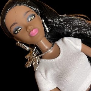 May include: A black doll with long black hair wearing a white t-shirt, a silver necklace, and silver earrings. The doll has brown skin, blue eyeshadow, and pink lips.