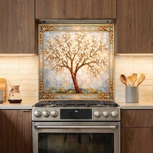 May include: A square wall art piece featuring a tree with white blossoms and a brown trunk, set against a mosaic-style background. The artwork is enclosed in an ornate, metallic gold frame with decorative corner details, positioned above a stove.