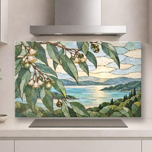 Puede incluir: Un panel rectangular para salpicadero de cocina con una ilustración estilo vidriera de ramas de eucalipto con hojas verdes y flores color crema en primer plano, sobre un paisaje costero con agua azul y colinas.