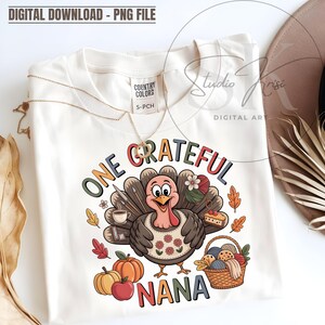Può includere: T-shirt bianco sporco con una grafica colorata di un tacchino. Il tacchino indossa un grembiule floreale e tiene forniture artistiche, lana e una fetta di torta. Il testo recita "ONE GRATEFUL NANA". Una collana d'oro è appoggiata sulla maglietta.