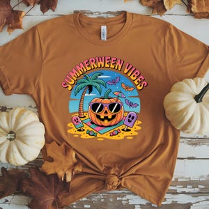 May include: Mustard-colored t-shirt with a colorful graphic that reads "Summerween Vibes." The graphic features a smiling pumpkin wearing sunglasses, a palm tree, bats, and ghost-shaped popsicles. The shirt is a crew neck.