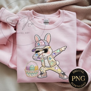 May include: A pale pink sweatshirt with a cartoon Easter bunny dabbing, wearing sunglasses, a hat, and a jacket. The bunny is holding a basket of Easter eggs. The words "Studio Krisi DIGITAL ART" are visible.
