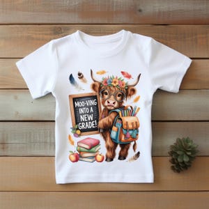 May include: White t-shirt featuring a cartoon illustration of a Highland cow wearing glasses and a flower crown, carrying a backpack and holding a chalkboard. The chalkboard reads "Moo-ving into a new grade!" with books and apples.