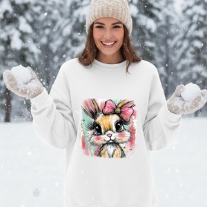 May include: White sweatshirt featuring a colorful illustration of a bunny with a pink bow. The bunny has large eyes and long eyelashes. The background is a snowy scene.