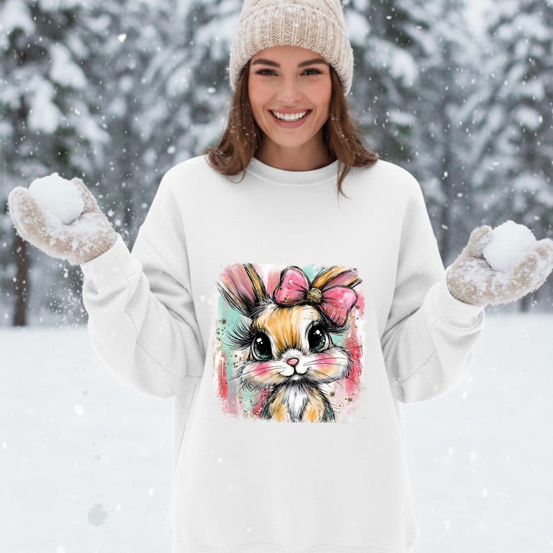 May include: White sweatshirt featuring a colorful illustration of a bunny with a pink bow. The bunny has large eyes and long eyelashes. The background is a snowy scene.