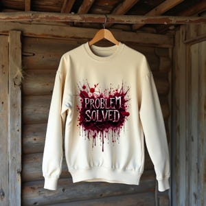 May include: Cream-colored sweatshirt with the text "PROBLEM SOLVED" in a dark red, splatter-style design. The crew neck sweatshirt is hanging on a wooden hanger against a rustic wooden background.