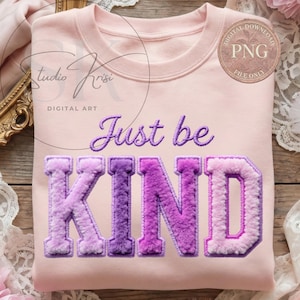 May include: A light pink sweatshirt with the embroidered phrase "Just be KIND" in purple. The word "KIND" is in a fuzzy, varsity-style font, with a gradient from light to dark purple. The sweatshirt has a crew neck and long sleeves.