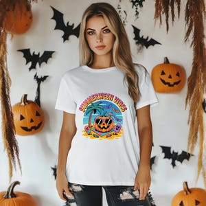 May include: White t-shirt with a colorful graphic that reads "Summerween Vibes." The graphic features a pumpkin wearing sunglasses on a beach with a palm tree, bats, and a ghost. Halloween-themed decorations are in the background.