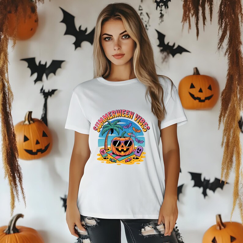 May include: White t-shirt with a colorful graphic that reads "Summerween Vibes." The graphic features a pumpkin wearing sunglasses on a beach with a palm tree, bats, and a ghost. Halloween-themed decorations are in the background.