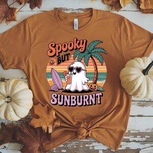 May include: Mustard-colored t-shirt with a graphic that reads "Spooky but Sunburnt." The design features a ghost wearing heart-shaped sunglasses, a cocktail, a surfboard, and a palm tree on a beach scene. Pumpkins and seashells are also included in the design.