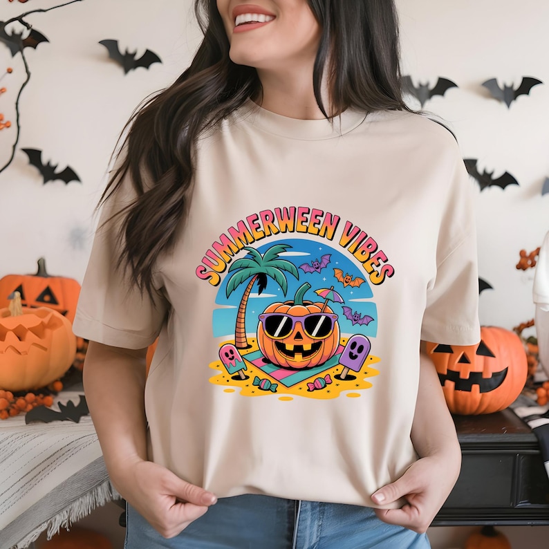 May include: Beige t-shirt with a colorful graphic that says "Summerween Vibes." The graphic features a pumpkin wearing sunglasses on a beach with bats, palm trees, and ghosts. Halloween and summer theme.