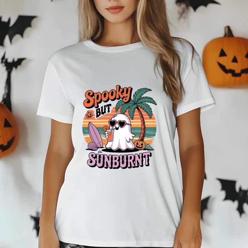May include: White t-shirt featuring a cartoon ghost wearing heart-shaped sunglasses, holding a drink, and enjoying a beach scene. The shirt has the words "Spooky but Sunburnt" in a colorful design.
