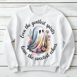 Peut inclure: Sweat-shirt blanc avec un motif de fantôme fantaisiste, des éléments floraux et le texte "Even the gentlest spirits leave the sweetest echoes." Le fantôme est dans des tons pastel. Vêtement confortable et stylé.