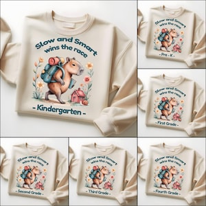 May include: Beige sweatshirt with a capybara illustration carrying a backpack, with the text "Slow and Smart wins the race" and "Kindergarten". Other sweatshirts show the same design with "Pre-K", "First Grade", "Second Grade", "Third Grade", and "Fourth Grade".