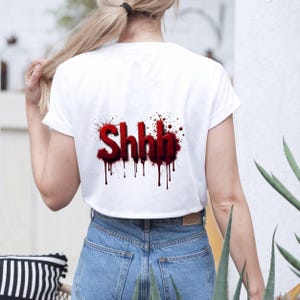 Pu&ograve; includere: Top corto bianco con la scritta "Shhh" in rosso, con un design a gocce simile a sangue. Il testo &egrave; stilizzato con un effetto 3D, e la maglietta &egrave; abbinata a jeans blu. L'estetica generale &egrave; audace e decisa.