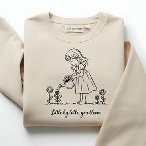 May include: A beige sweatshirt with a black illustration of a girl watering flowers. The text "Little by little, you bloom" is below the illustration. The sweatshirt has a Luxe Comfort label.