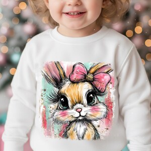 May include: White long-sleeved sweatshirt featuring a colorful illustration of a bunny with a pink bow. The bunny has large eyes and long eyelashes. The design is set against a backdrop of pink, teal, and gold accents.