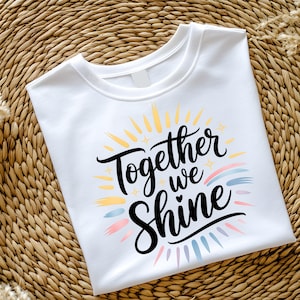 Może przedstawiać: Biała koszulka z okrągłym dekoltem z napisem "Together we Shine" w czarnym skrypcie. Projekt zawiera kolorowe akcenty promieni słonecznych i tęczy w kolorze żółtym, niebieskim, różowym i fioletowym. Koszulka jest złożona na tkanej powierzchni.