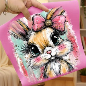 May include: A pink square featuring a cartoon bunny with large, expressive eyes and long eyelashes. The bunny has a pink bow with a gold center and is set against a colorful, abstract background with gold accents. The bunny has a pink nose and blush.