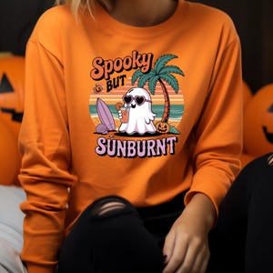 May include: Orange sweatshirt with a graphic of a ghost wearing heart-shaped sunglasses, holding a drink, and standing on a beach with a palm tree. The text reads "Spooky but Sunburnt".