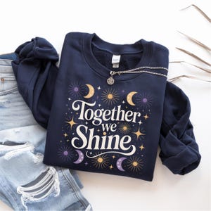Puede incluir: Una sudadera azul marino con el texto "Together we Shine" en escritura blanca. El diseño incluye estrellas, lunas y rayos de sol dorados y morados. Un collar plateado con un colgante descansa sobre la sudadera. La sudadera está doblada junto a unos vaqueros azules.