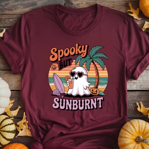May include: Burgundy t-shirt with a graphic of a ghost wearing heart-shaped sunglasses, holding a drink, and relaxing on a beach. The text reads "Spooky but Sunburnt" in orange and purple. A palm tree, surfboard, and pumpkins are also featured.
