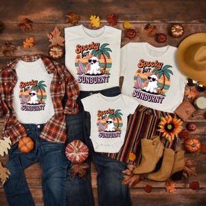 May include: A collection of white t-shirts and a baby onesie with a Halloween-themed graphic. The design features a ghost wearing sunglasses, a surfboard, and the text "Spooky But Sunburnt". The shirts are paired with jeans, a flannel shirt, and boots.