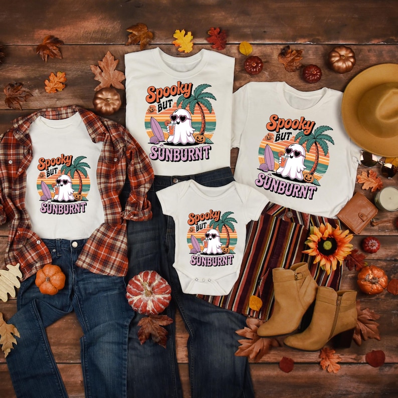 May include: A collection of white t-shirts and a baby onesie with a Halloween-themed graphic. The design features a ghost wearing sunglasses, a surfboard, and the text "Spooky But Sunburnt". The shirts are paired with jeans, a flannel shirt, and boots.