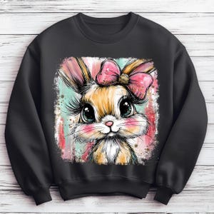 May include: Black sweatshirt featuring a colorful illustration of a bunny with a pink bow. The bunny has large eyes, long eyelashes, and pink cheeks. The background has vertical stripes in shades of pink, blue, and white.