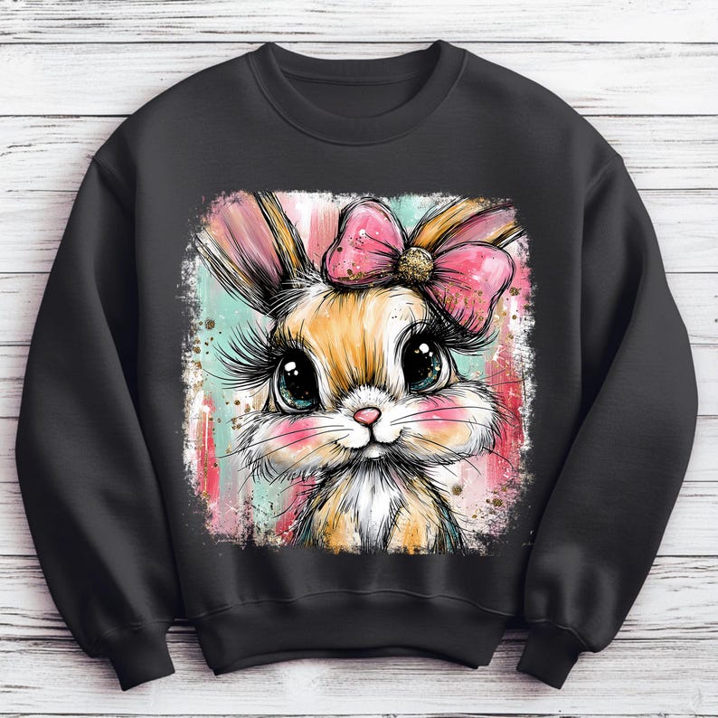 May include: Black sweatshirt featuring a colorful illustration of a bunny with a pink bow. The bunny has large eyes, long eyelashes, and pink cheeks. The background has vertical stripes in shades of pink, blue, and white.