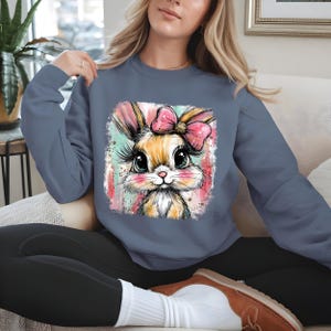 May include: A blue sweatshirt featuring a colorful graphic of a bunny with a pink bow. The bunny has large eyes and long eyelashes. The graphic is set against a watercolor-style background with pink and teal accents. The sweatshirt is a solid blue color.