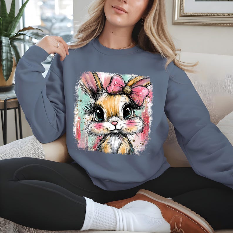 May include: A blue sweatshirt featuring a colorful graphic of a bunny with a pink bow. The bunny has large eyes and long eyelashes. The graphic is set against a watercolor-style background with pink and teal accents. The sweatshirt is a solid blue color.