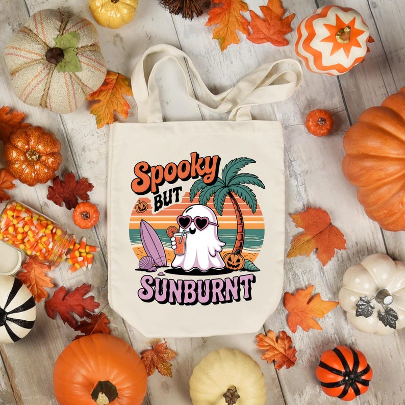 May include: A cream-colored tote bag with the words "Spooky but Sunburnt" in orange and purple. The graphic features a ghost wearing sunglasses, holding a drink, and a surfboard on a beach with a palm tree. Pumpkins and fall leaves surround the bag.