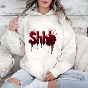 Pu&ograve; includere: Felpa con cappuccio color crema con la scritta "Shhh" in lettere rosse con effetto gocciolante di sangue. La grafica &egrave; circondata da schizzi rossi. La felpa ha una tasca frontale e un cappuccio con coulisse.