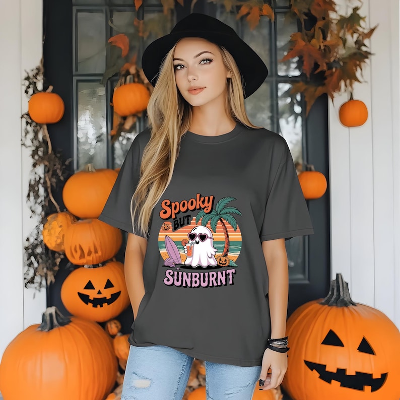 May include: Dark gray t-shirt with a graphic that reads "Spooky But Sunburnt". The design features a ghost wearing sunglasses, a surfboard, and a palm tree. The shirt is part of a Halloween-themed collection.