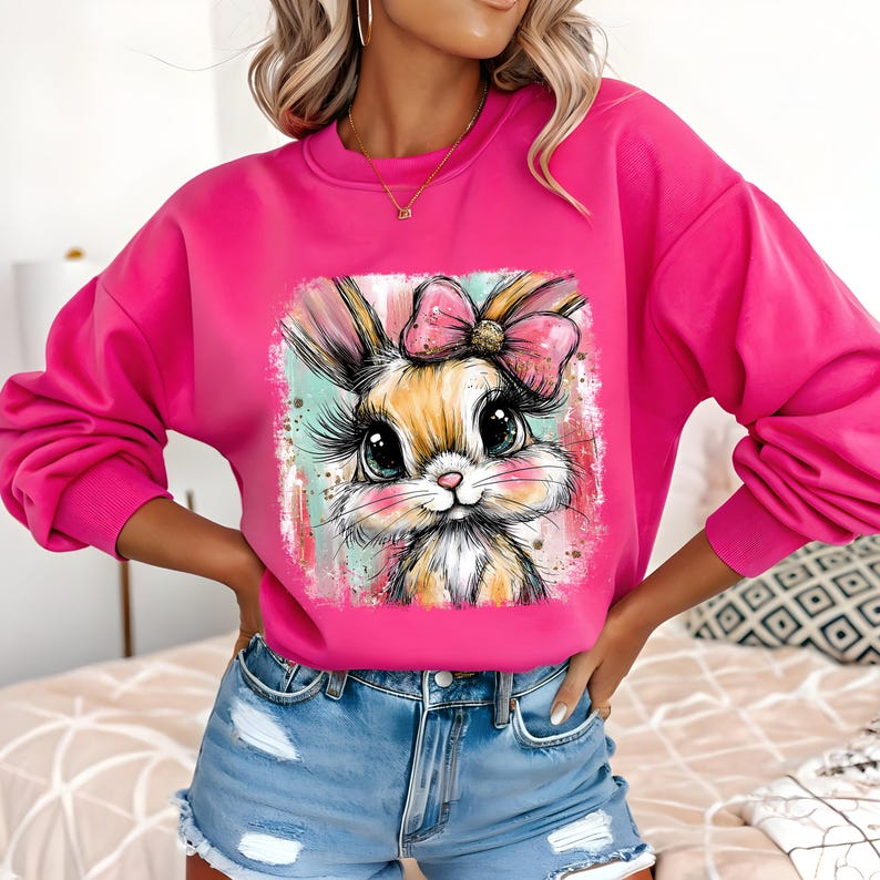 May include: A bright pink sweatshirt featuring a colorful graphic of a bunny with a pink bow. The bunny has large eyes and long eyelashes. The sweatshirt has long sleeves and a crew neck.