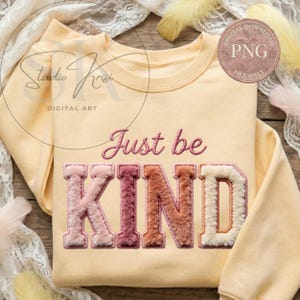 Può includere: Una felpa giallo chiaro con la scritta ricamata "Just be KIND" in rosa e varie sfumature di rosa, marrone e crema. La felpa ha un girocollo e maniche lunghe. Sono visibili anche le parole "Studio Krisi" e "Digital Art".