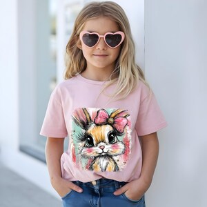 May include: A light pink t-shirt featuring a colorful illustration of a bunny with a pink bow. The bunny has large eyes and long eyelashes. The girl is wearing pink heart-shaped sunglasses and blue jeans.