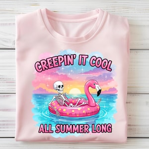 Op de afbeelding: Lichtroze T-shirt met een afbeelding van een skelet op een roze flamingo-opblaasboot in het water. De tekst "CREEPIN' IT COOL ALL SUMMER LONG" staat boven en onder de afbeelding, tegen een achtergrond van zonsondergang.