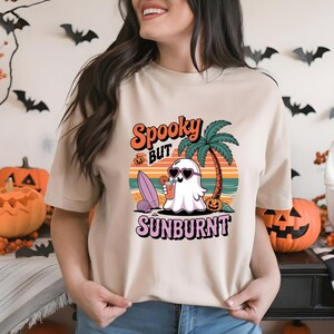 May include: Beige t-shirt with a graphic featuring a ghost wearing heart-shaped sunglasses, holding a drink, and a palm tree. The text reads "Spooky but Sunburnt" in orange and pink. A surfboard and pumpkin are also in the design.